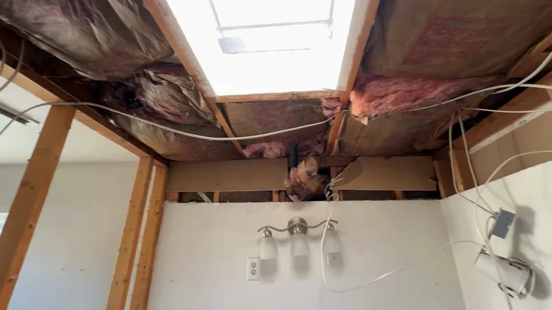 A bathroom ceiling opened up to bare joists and pink insulation, with a skylight in the middle