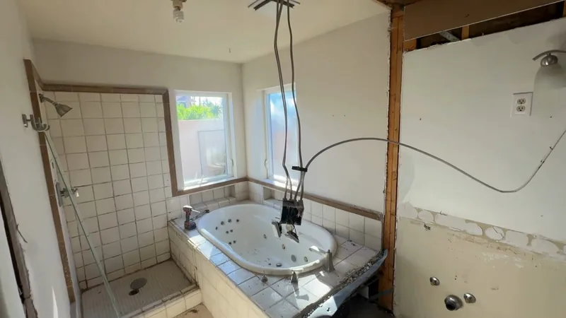 A bathroom stripped to its studs and joists around a tiled tub and shower, with wires hanging from the ceiling