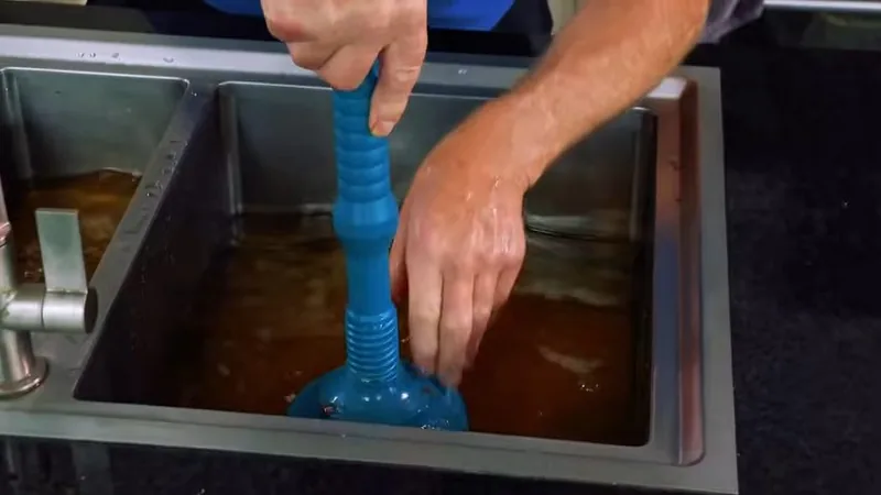 Hands working a blue plunger in a steel sink that holds murky standing water