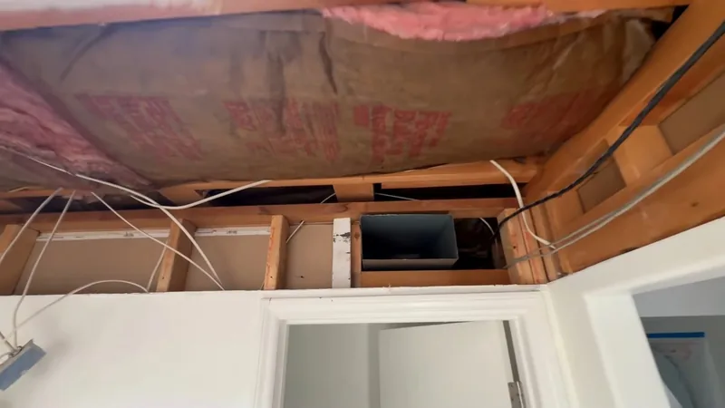 An opened ceiling showing pink insulation, wood joists and wiring above a bathroom doorway