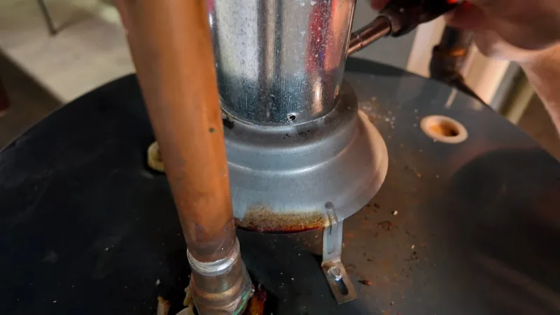 A copper pipe beside a metal vent pipe on top of a black water heater, with a hand and a torch at the top right