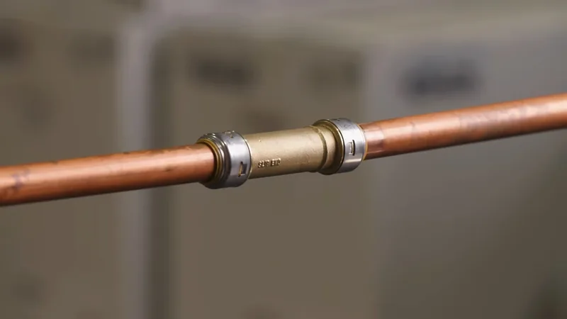 A straight copper pipe joined by a metal coupling with a blurred wall behind it