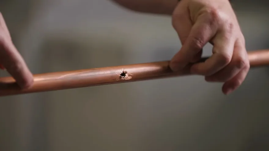 Fingers holding a straight length of copper pipe with a small split hole in its wall