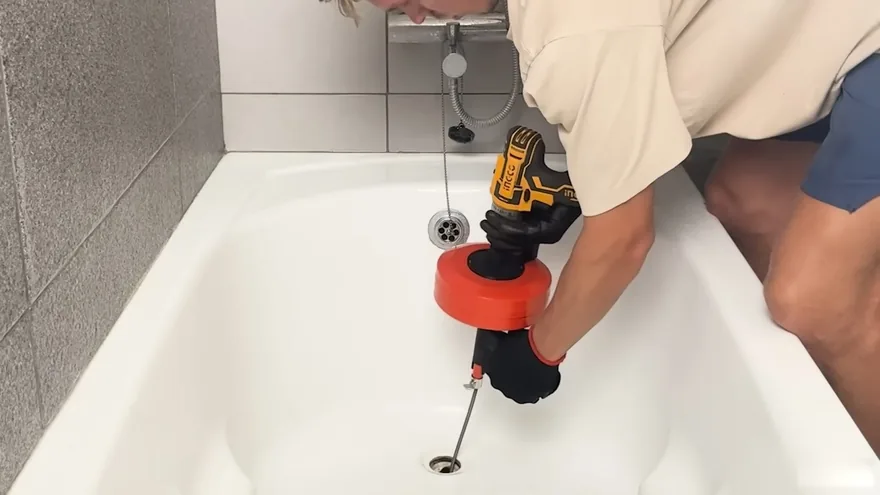 A person using a cordless drill with a red drain auger drum to clear a bathtub drain
