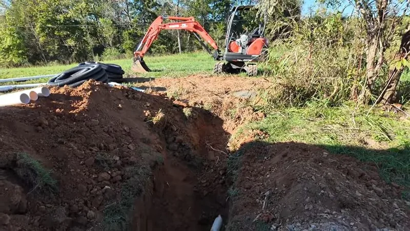 A small orange excavator beside an open dirt trench with white pipe ends and coils of black pipe on the ground