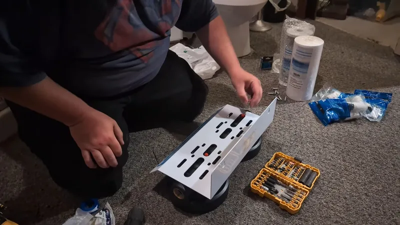 A person kneeling on carpet beside a white mounting bracket, drill bits and two sediment filter cartridges