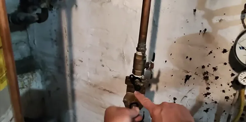 A finger pointing at a brass valve on a copper pipe beside a pressure gauge on a spattered wall