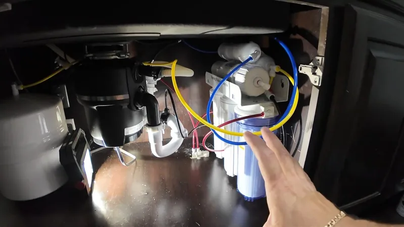 The inside of a dark sink cabinet with a garbage disposal on the left, a white drain trap and a blue filter canister on the right, and a hand gesturing toward them