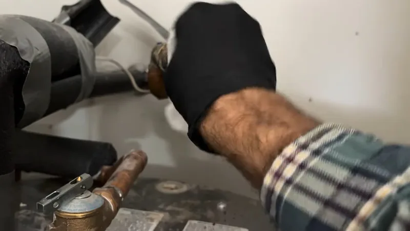 A gloved hand adjusting a brass fitting on the inlet side of a water heater beside insulated pipe