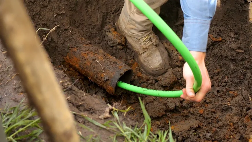 A worker in boots feeding a green flexible line into the open end of a rusty pipe in a dirt trench