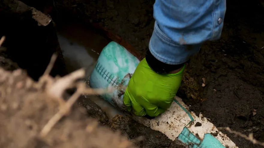 A green-gloved hand pressing a teal liner sleeve in a dirt trench around a white pipe