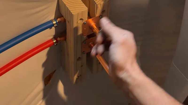 A hand at copper fittings mounted on a wood block with a blue and a red supply line running to them