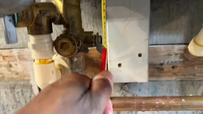 A hand beside a brass shutoff valve and white fittings on a wall, with a tape measure hanging next to it