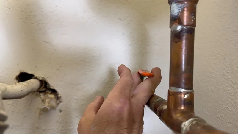 A hand beside a copper elbow on a wall, with a small broken opening in the plaster and a white pipe beside it