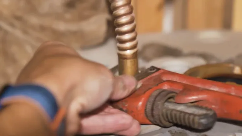 A hand holding a red pipe wrench on a copper line above a water heater