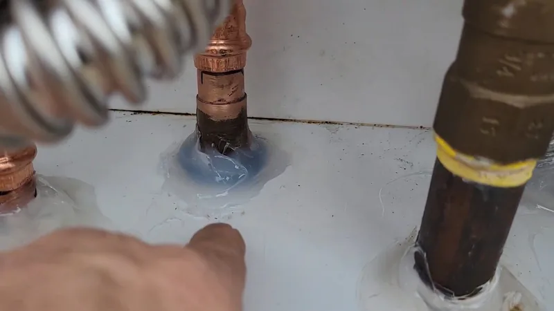 A hand near a copper pipe sealed with blue compound, a flexible metal line and a brass pipe on a white surface