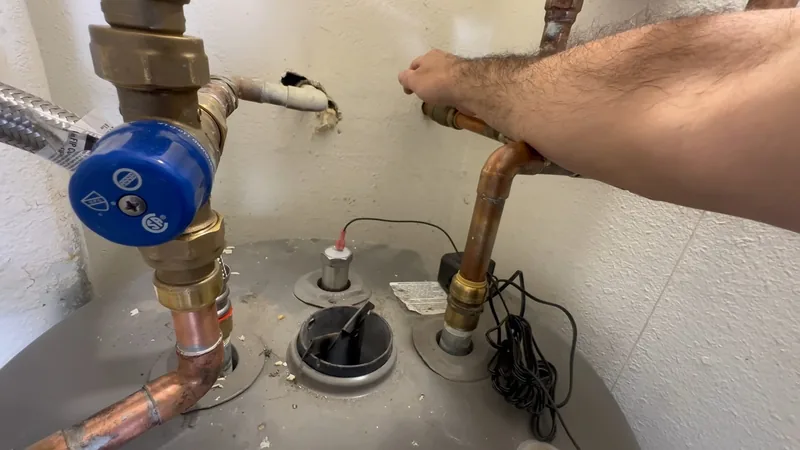 A hand on a copper pipe connection on top of a water heater beside a brass fitting with a blue handle