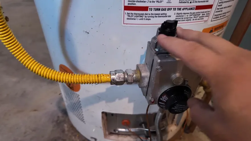 A hand on the gas control of a white water heater with a yellow flexible gas line attached