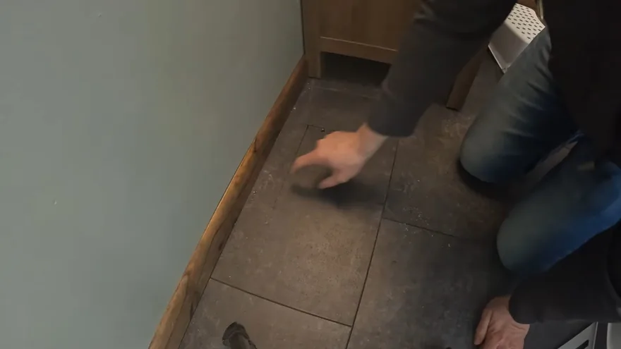 A person kneeling beside a baseboard with one hand pressed flat on a dark tile floor