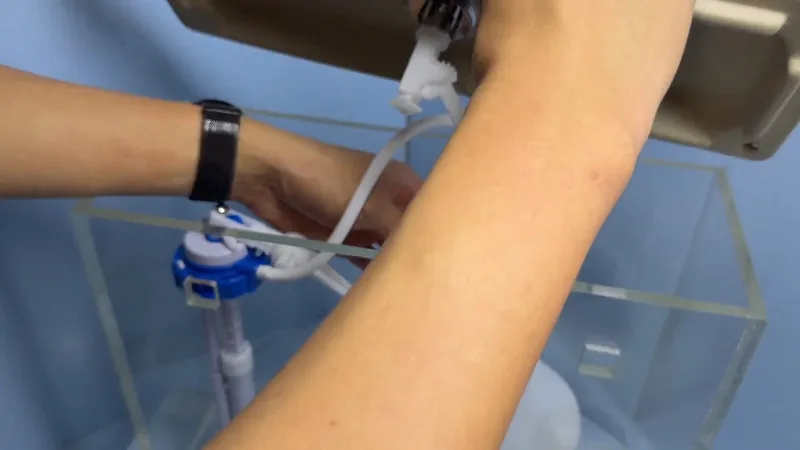Hands adjusting a blue and white fill valve inside a clear acrylic toilet tank