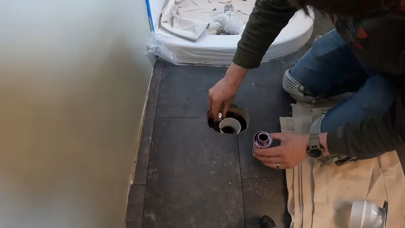 Hands holding a can of purple primer beside an open toilet flange in a dark floor