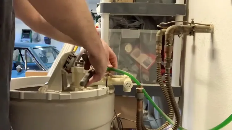 Hands connecting a green hose to the valve on top of a cream-colored softener tank beside brass pipes