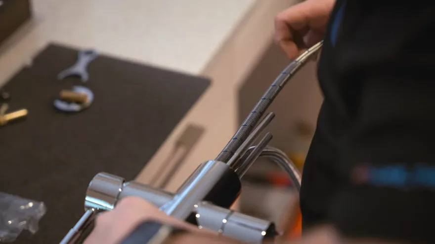 Hands holding a chrome faucet body with a braided supply hose beside wrenches on a dark mat
