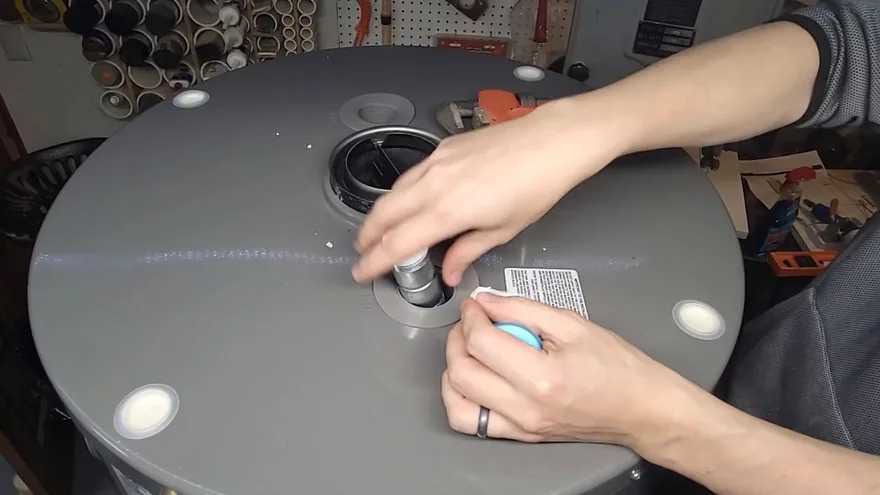 Two hands tightening a threaded fitting on the top of a gray tank water heater in a workshop