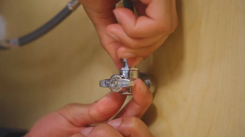 Hands turning a small chrome shutoff valve with a braided supply line against a wood panel
