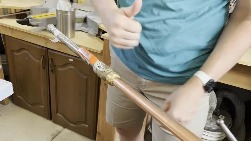 A man in a teal shirt holding a length of copper pipe with a flexible connector over a workbench
