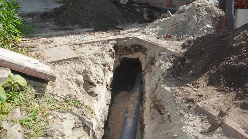 A narrow excavation beside a slab with a dark sewer pipe exposed at the bottom