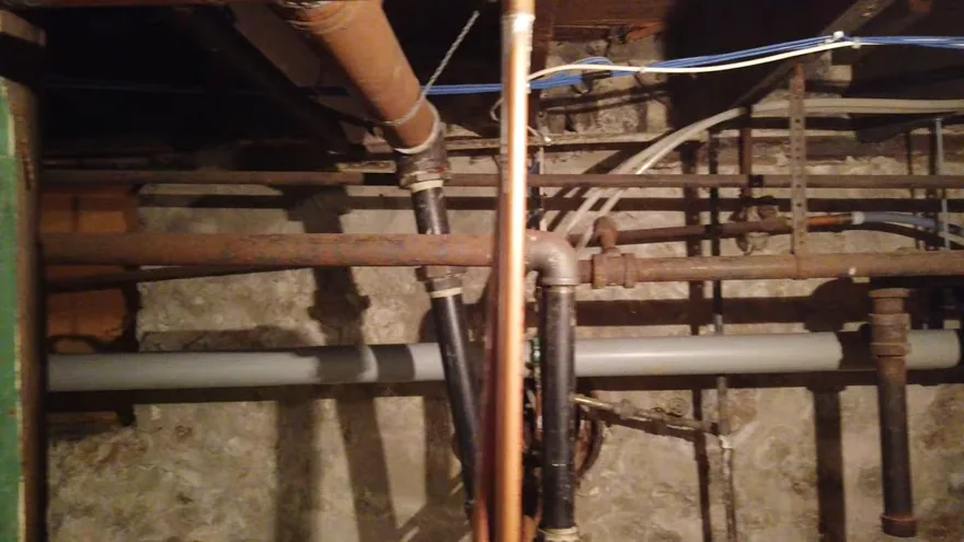 Old rusted steel, copper and gray plastic pipes running between joists in a crawl space
