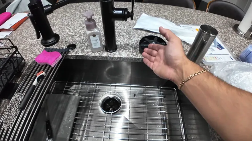 An open hand held over a stainless steel sink with a drain flange, a rack and a black faucet