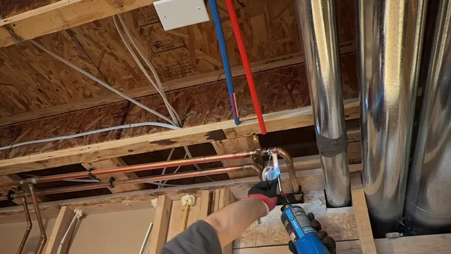 A gloved hand holding a torch to a copper joint between ceiling joists, with red and blue PEX lines above