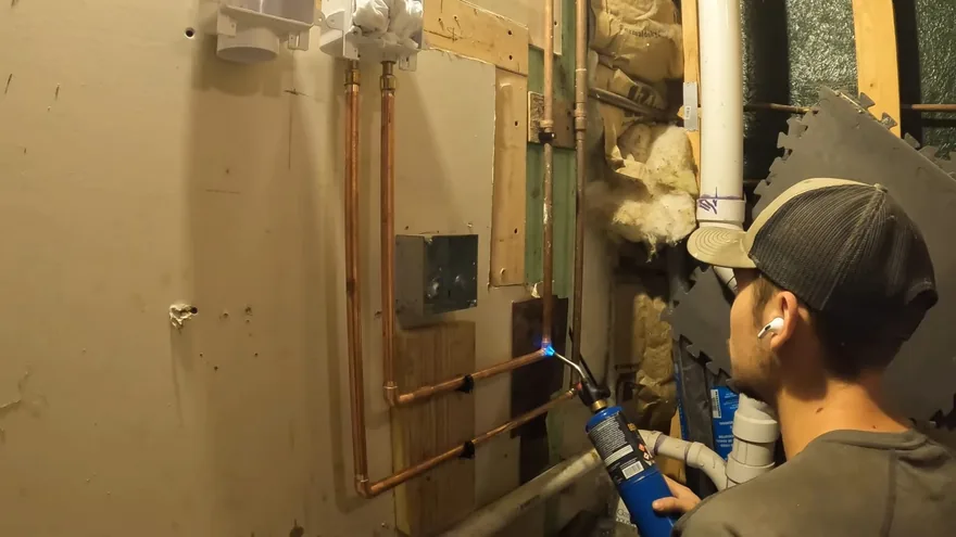 A plumber in a cap holding a blue torch to copper supply pipes on a utility room wall