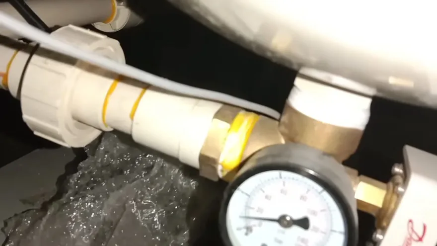 A round pressure gauge threaded into brass and white plastic pipe fittings beside a tank wrapped in plastic