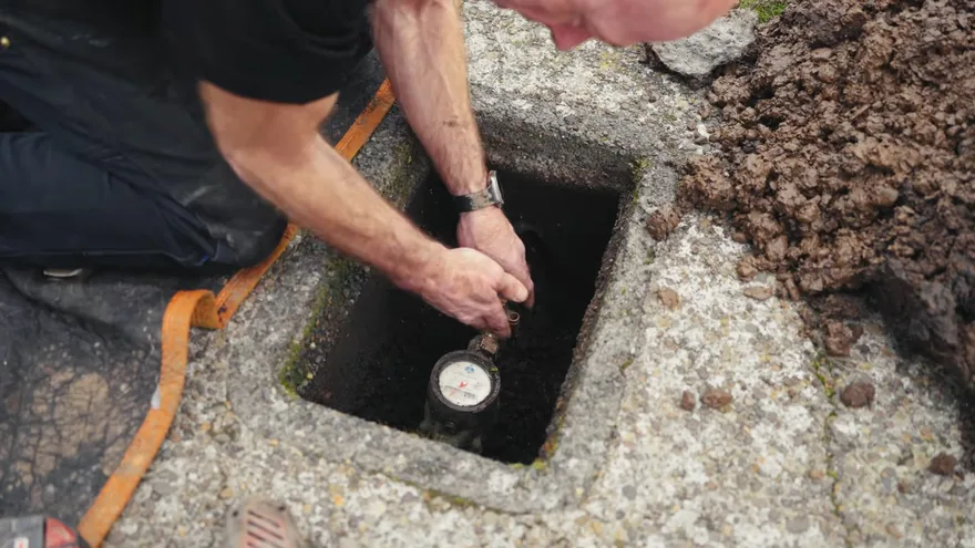 Hands steadying a water meter inside an in-ground meter box beside a pile of soil
