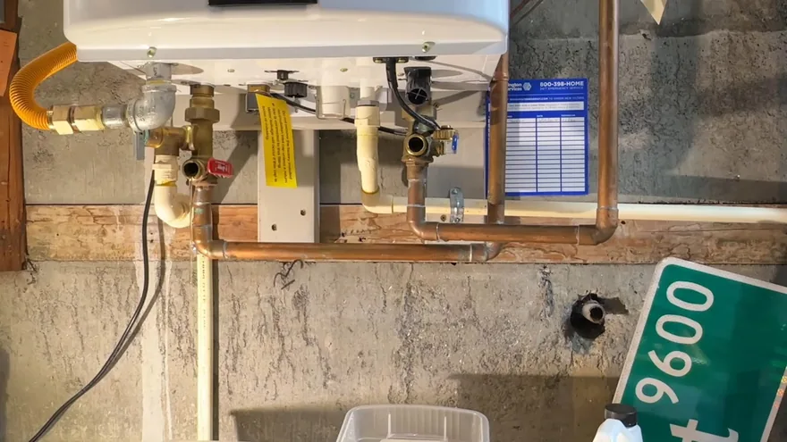 A white tankless water heater on a concrete wall with copper pipes, service valves and a yellow gas tag