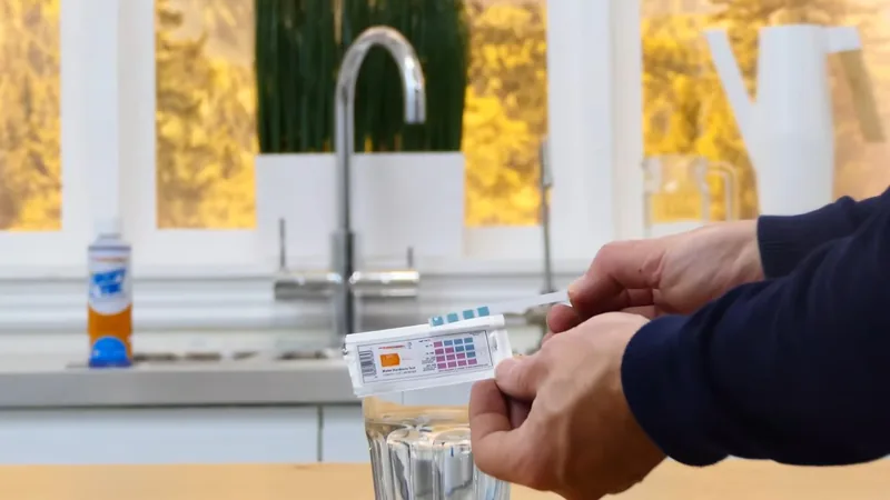 Hands holding a test strip card over a glass of water beside a kitchen faucet