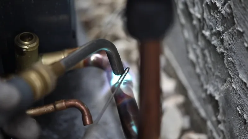 A torch flame heating a copper pipe fitting beside a rough stucco wall