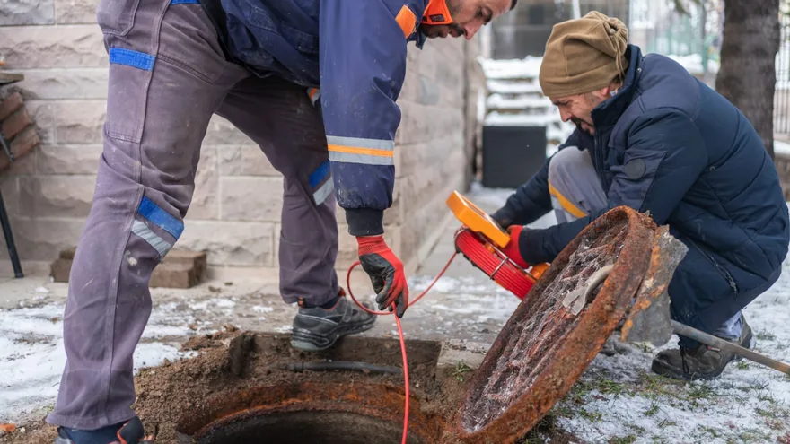 Two workers beside an opened manhole with a red camera cable and an orange reel on snowy ground