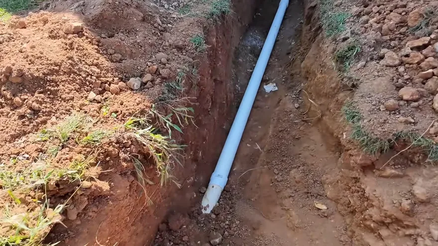 A white plastic pipe lying in the bottom of a narrow dirt trench with grass along its edges
